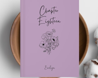 May include: A lavender-colored book with the words "Chapter Eighteen" in black script. Below the text is a black and white floral illustration, and the name "Evelyn" is written at the bottom. The book is resting in a wooden bowl.