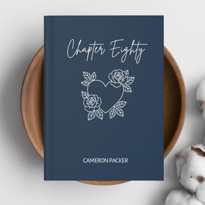 Puede incluir: Un libro azul marino con el título "Chapter Eighty" en escritura blanca. Un diseño de corazón floral blanco está centrado en la portada, con el nombre del autor, "CAMERON PACKER", debajo. El libro está en un cuenco de madera.