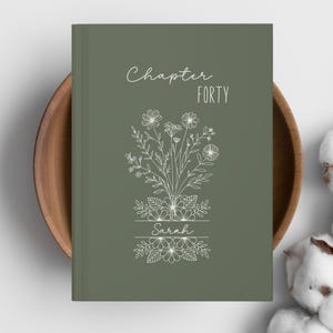 May include: A sage green journal with the words "Chapter Forty" and "Sarah" in white script. A detailed floral illustration is centered on the cover. The journal is resting in a wooden bowl.