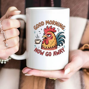 Puede incluir: Taza de cerámica blanca con un diseño de gallo de dibujos animados, sosteniendo una taza de café y con una expresión de mal humor. La taza tiene el texto "GOOD MORNING NOW GO AWAY". El gallo es rojo, amarillo y azul.