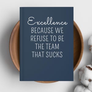 May include: A navy blue notebook with the words "Excellence BECAUSE WE REFUSE TO BE THE TEAM THAT SUCKS" in white script. The notebook is in a wooden bowl, with cotton buds in the background.