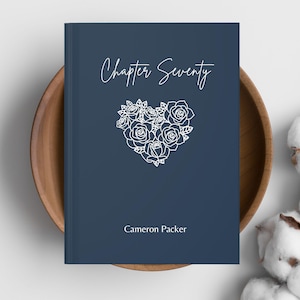 May include: A navy blue book with the title "Chapter Seventy" in cursive script. A white floral heart design is centered on the cover, with the author's name, Cameron Packer, below. The book is resting in a wooden bowl.