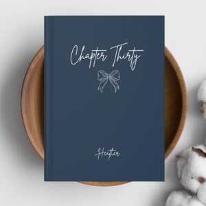 May include: A navy blue journal with the words "Chapter Thirty" and a bow illustration in white script. The name "Heather" is written at the bottom. The journal is in a wooden bowl.