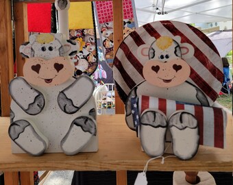 以下が含まれることがあります： 白黒の牛柄に塗られた木製の牛の置物2つ。1つの牛は小さなアメリカ国旗を持ち、もう1つの牛は赤白青のストライプのペーパータオルホルダーを持っている。