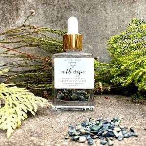 May include: A clear glass bottle with a gold lid and dropper filled with green and brown crystals. The bottle label reads "RITUAL:WILD earth magic element ritual oil (gemstone-infused) cedarwood aged dark patchouli".