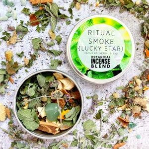 May include: A round metal tin with a green lid containing a blend of dried herbs and spices. The lid has a label that reads "Ritual Smoke {Lucky Star} Prosperity Herbal Blend Botanical Incense Blend".