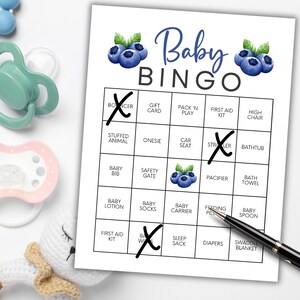 Peut inclure: Une carte de jeu Baby Bingo avec des illustrations de myrtilles et les mots "Baby Bingo" en &eacute;criture bleue. La carte comporte des cases avec des termes li&eacute;s aux b&eacute;b&eacute;s, dont certains sont marqu&eacute;s d'un "X". Des sucettes, un jouet pour b&eacute;b&eacute; et un stylo sont &eacute;galement visibles.