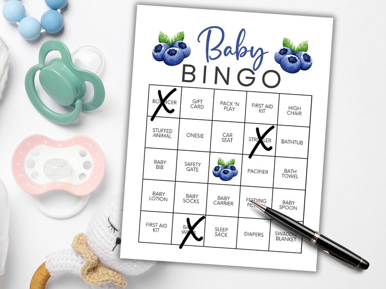 Peut inclure: Une carte de jeu Baby Bingo avec des illustrations de myrtilles et les mots "Baby Bingo" en &eacute;criture bleue. La carte comporte des cases avec des termes li&eacute;s aux b&eacute;b&eacute;s, dont certains sont marqu&eacute;s d'un "X". Des sucettes, un jouet pour b&eacute;b&eacute; et un stylo sont &eacute;galement visibles.