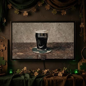 May include: A television screen displays a painting of a pint of Guinness stout on a coaster. The dark beer contrasts with the white head, set against a neutral background. The scene is decorated with shamrock garlands and green accents, evoking an Irish pub atmosphere.