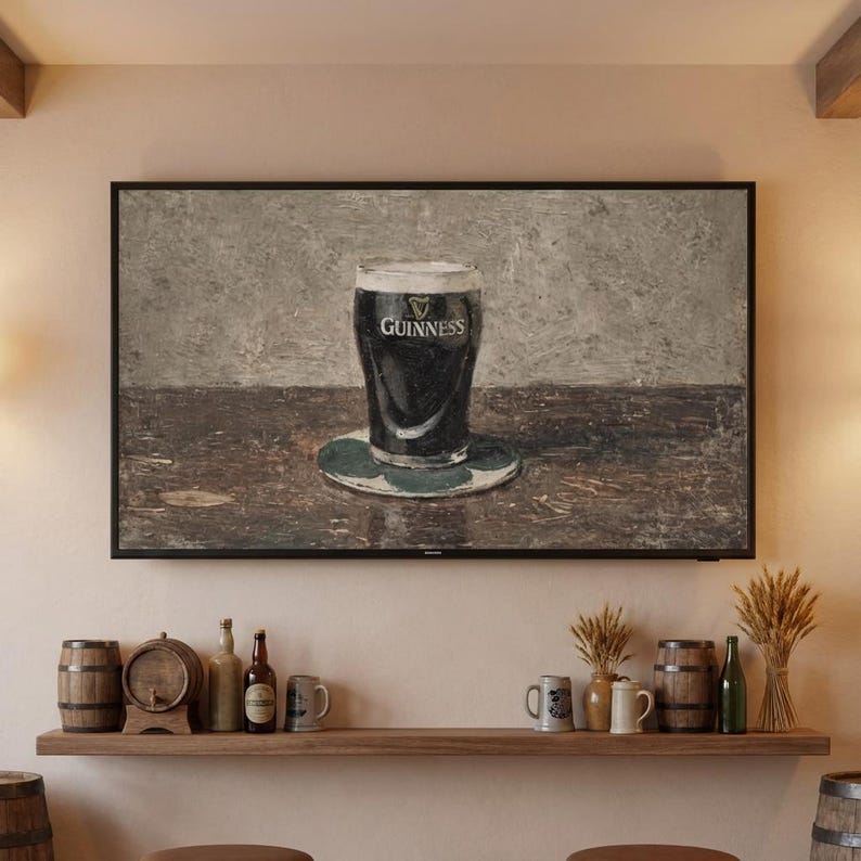 May include: A framed print of a pint of Guinness stout on a green coaster. The glass is filled with dark liquid and topped with a creamy head. The Guinness logo is visible. The print is displayed above a wooden shelf with bar accessories.
