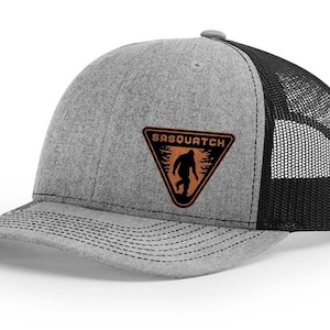 May include: Gray trucker hat with a black mesh back and a curved brim. The front features a brown triangle patch with the word "SASQUATCH" above a silhouette of the creature.