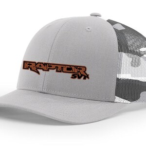 May include: A gray and white camouflage trucker hat with a brown embroidered logo that says "RAPTOR SVT".