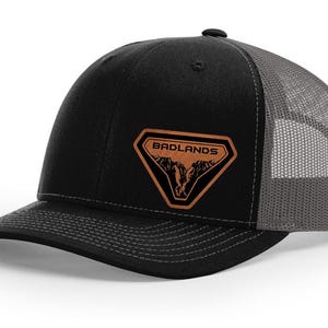 May include: Black trucker cap with a grey mesh back and a curved peak. It has a brown leather patch on the front with the word "BADLANDS" above a mountain graphic. The cap has a snapback fastening.