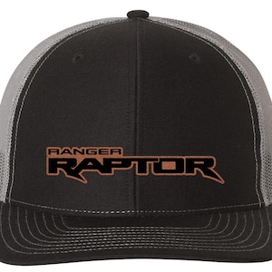 May include: Black and gray trucker hat with the words "RANGER RAPTOR" in brown lettering. The hat has a curved brim and a mesh back for breathability. The hat is a classic style.