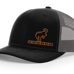May include: Black and grey trucker hat with a brown leather patch featuring the Ford Bronco logo. The logo is a silhouette of a rearing horse with the word "Bronco" underneath.