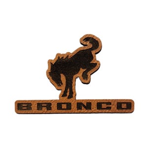 May include: A brown and black Bronco logo patch. The patch features a bucking horse silhouette above the word "BRONCO" in capital letters. The patch is made of a textured material.