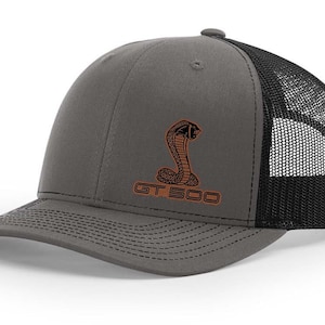 May include: A gray and black trucker hat with a black mesh back. The front of the hat features an orange embroidered cobra logo and the text "GT 500".
