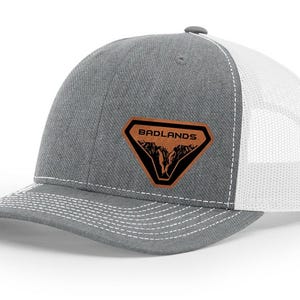 May include: A grey and white trucker cap with a curved peak. The front panel is grey with a brown and black patch that reads "BADLANDS" above a mountain graphic. The back is white mesh.