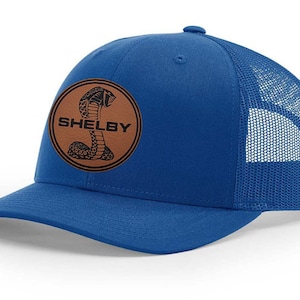 Peut inclure: Un chapeau de camionneur bleu royal avec un patch en cuir marron représentant un cobra noir et le mot "SHELBY" en blanc.