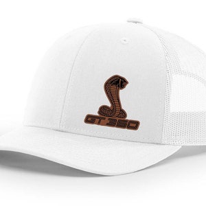 Peut inclure: Casquette de camionneur blanche avec un patch en cuir marron représentant un cobra noir et le texte "GT 350".