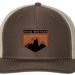 May include: A brown and tan trucker hat with a Big Bend patch. The patch features a mountain range and the words "BIG BEND" in a rectangular frame. The hat has a curved brim and a mesh back.