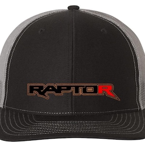 May include: Black and gray trucker hat with the word "RAPTOR" in orange and red across the front. The hat has a curved brim and a mesh back for breathability. The hat is a classic style.