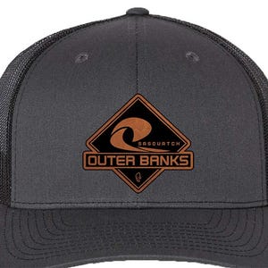 May include: A charcoal gray trucker hat with a black mesh back. The front features a brown and black patch with the words "SASQUATCH" and "OUTER BANKS" and a wave design.