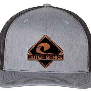 May include: Gray trucker hat with a black mesh back and a curved bill. The front features a brown and black diamond-shaped patch with the words "Outer Banks" and a wave design.