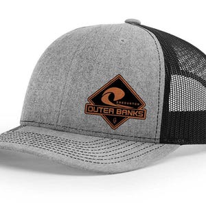May include: Gray trucker hat with a black mesh back and a curved brim. The front features a brown and black diamond-shaped patch with the text "Outer Banks" and a wave graphic.