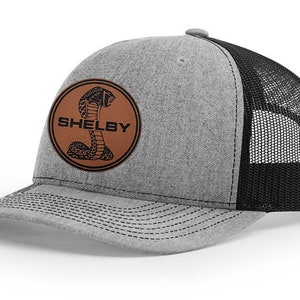 Peut inclure: Un chapeau de camionneur gris et noir avec un patch en cuir marron représentant un cobra noir et le mot "SHELBY".