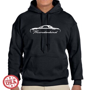May include: Black hooded sweatshirt with a white silhouette of a Ford Thunderbird and the text "Thunderbird" printed on the front. The sweatshirt has a kangaroo pocket and a drawstring hood. The tag on the sleeve reads "Made in USA."