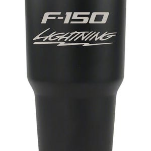 May include: A matte black stainless steel tumbler featuring a silver metallic rim at the top and bottom. The front of the cup displays the text F-150 in a bold font above the word Lightning in a stylized script font with a horizontal line underneath.