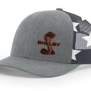 Peut inclure: Un chapeau de camionneur gris et bleu avec un motif d'étoiles blanches à l'arrière. L'avant du chapeau présente un logo brodé Shelby Cobra noir et orange avec le mot "SHELBY" en dessous.