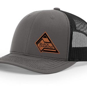 May include: A gray trucker hat with a black mesh back and a curved brim. The hat features a brown and black diamond-shaped patch with the words "BLACK DIAMOND" and a mountain graphic.