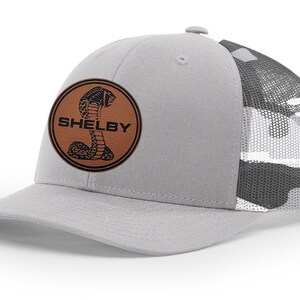 Peut inclure: Un chapeau de camionneur en camouflage gris et noir avec un patch en cuir marron représentant un cobra noir et le mot "SHELBY" en texte noir.