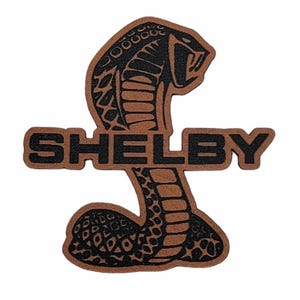 May include: A brown and black Shelby Cobra logo patch. The patch features a cobra snake with its mouth open, above the word "SHELBY" in bold, black letters. The patch is likely made of leather or a similar material.