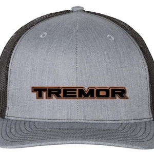 May include: A gray trucker hat with a black mesh back and a curved brim. The word "TREMOR" is embroidered in black with a brown outline across the front of the hat. The hat has a structured front panel and a snapback closure.