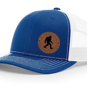 May include: A blue and white trucker hat with a brown leather patch featuring a silhouette of Bigfoot and the text "Bigfoot Research Team".