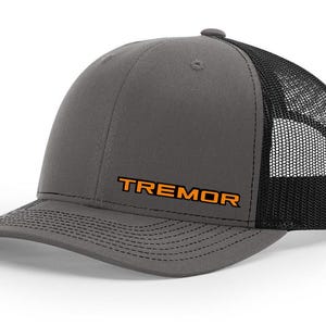 May include: A grey trucker cap with a black mesh back and a curved peak. The word "TREMOR" is printed in orange across the front. The cap has a snap closure for an adjustable fit.
