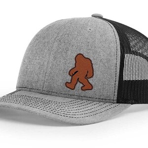 May include: A gray and black trucker hat with a brown felt Bigfoot silhouette patch.
