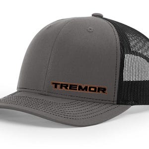May include: A grey trucker cap with a black mesh back. The word "TREMOR" is embroidered in orange on the front. The cap has a curved peak and a snapback fastening.