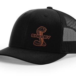 Peut inclure: Casquette de camionneur noire avec un logo Shelby brodé en marron représentant un cobra.