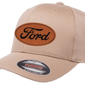 Puede incluir: Una gorra de béisbol de color beige con un parche ovalado de cuero marrón que dice "Ford" en letras negras. La gorra tiene una etiqueta Flexfit en la parte inferior de la visera.