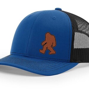 May include: A royal blue trucker hat with a black mesh back. The hat features a brown embroidered silhouette of Bigfoot.