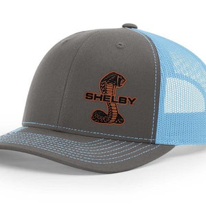 Peut inclure: Un chapeau de camionneur gris et bleu clair avec un logo brodé Shelby Cobra noir et orange. Le logo comporte le texte "Shelby" et un cobra stylisé.