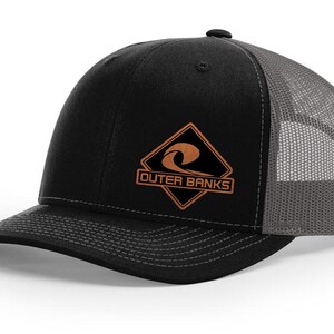 May include: Black and gray trucker hat with a curved brim. The front panel is black with a brown and orange diamond-shaped patch that reads "Outer Banks". The mesh back is gray.