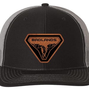 May include: Black and gray trucker hat with a brown leather patch. The patch features the word "BADLANDS" above a mountain graphic. The hat has a curved brim and a mesh back.