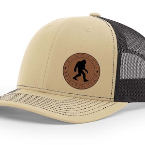May include: A beige and black trucker hat with a brown leather patch that says "Bigfoot Research Team" and has a silhouette of Bigfoot walking.