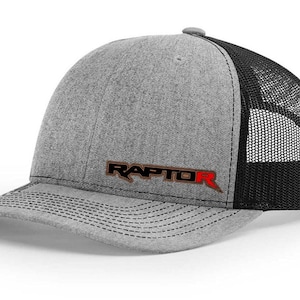 May include: A gray and black trucker hat with the word "RAPTOR" in brown and red lettering. The hat has a curved brim and a mesh back for ventilation. The hat is a baseball cap style.