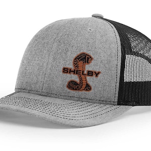 Peut inclure: Un chapeau de camionneur gris et noir avec un cobra brodé noir et le mot "SHELBY" en orange.